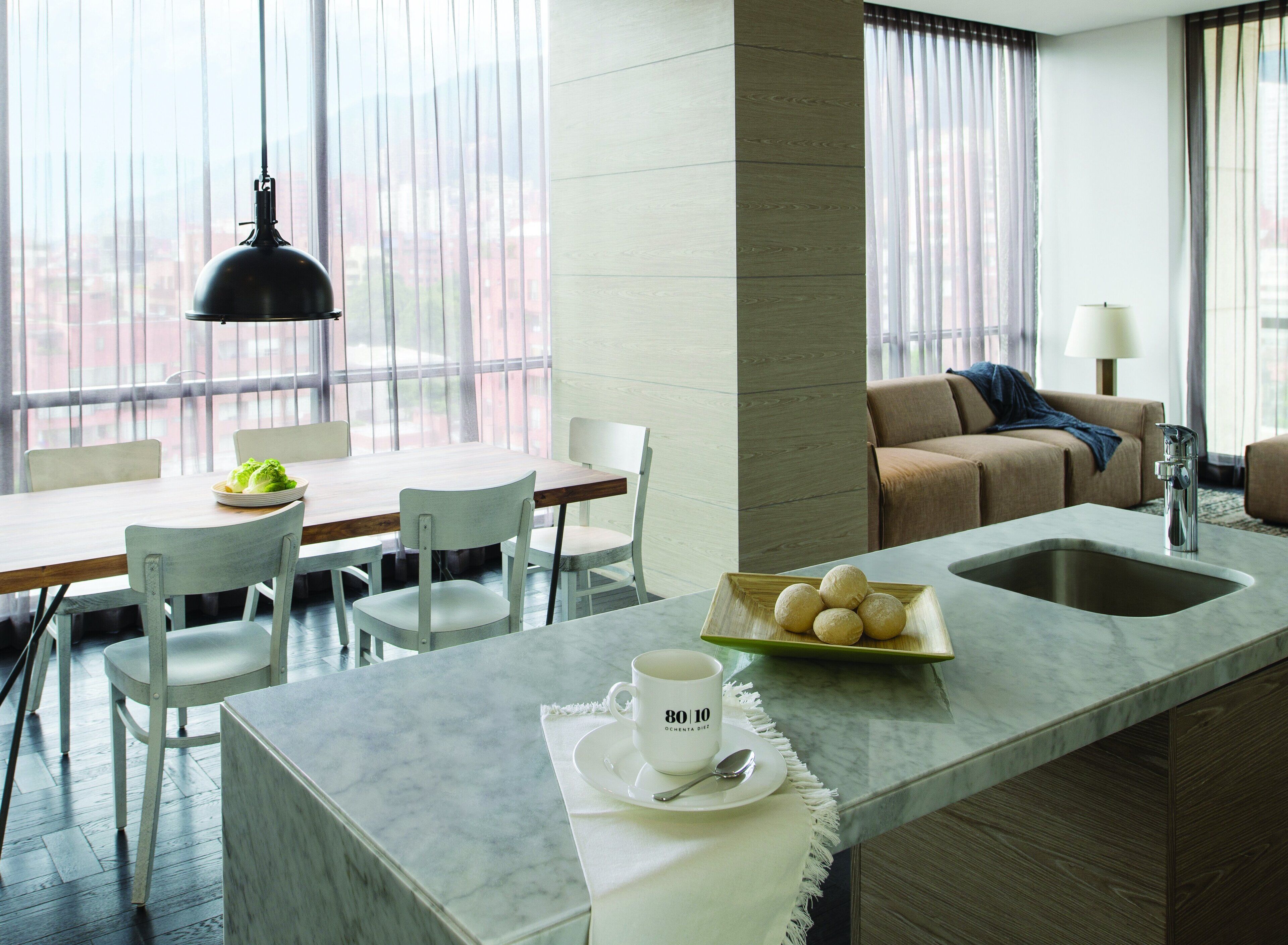 A modern kitchen with a marble countertop, a dining area with chairs, a sofa, and a view through large windows.