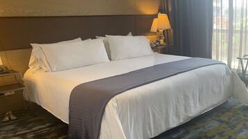 A hotel room with a large bed, a bedside table with a lamp, and a view of the outdoors through the window.