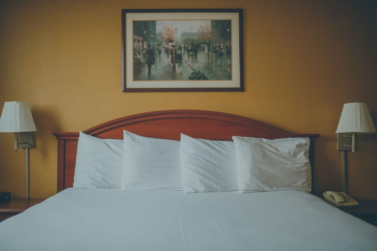 A hotel room with a large bed, white linens, a wooden headboard, two bedside lamps, a framed painting on the wall, and a bedside telephone.