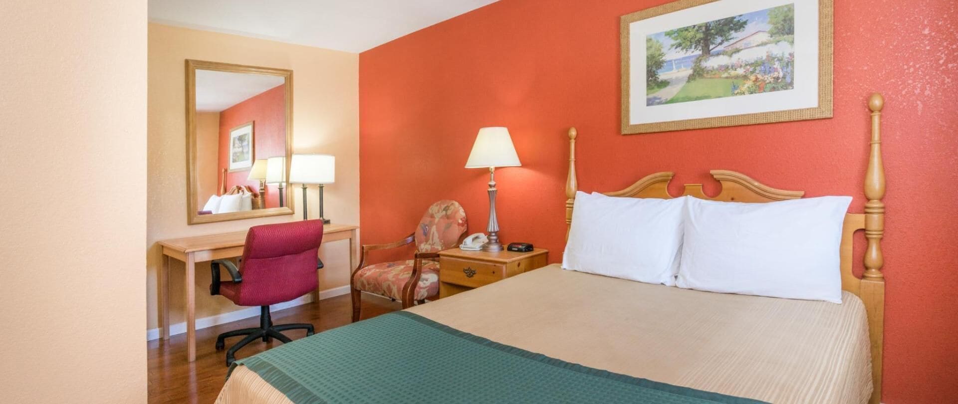 A hotel room with a bed, a desk, a chair, a lamp, and a painting on the wall.