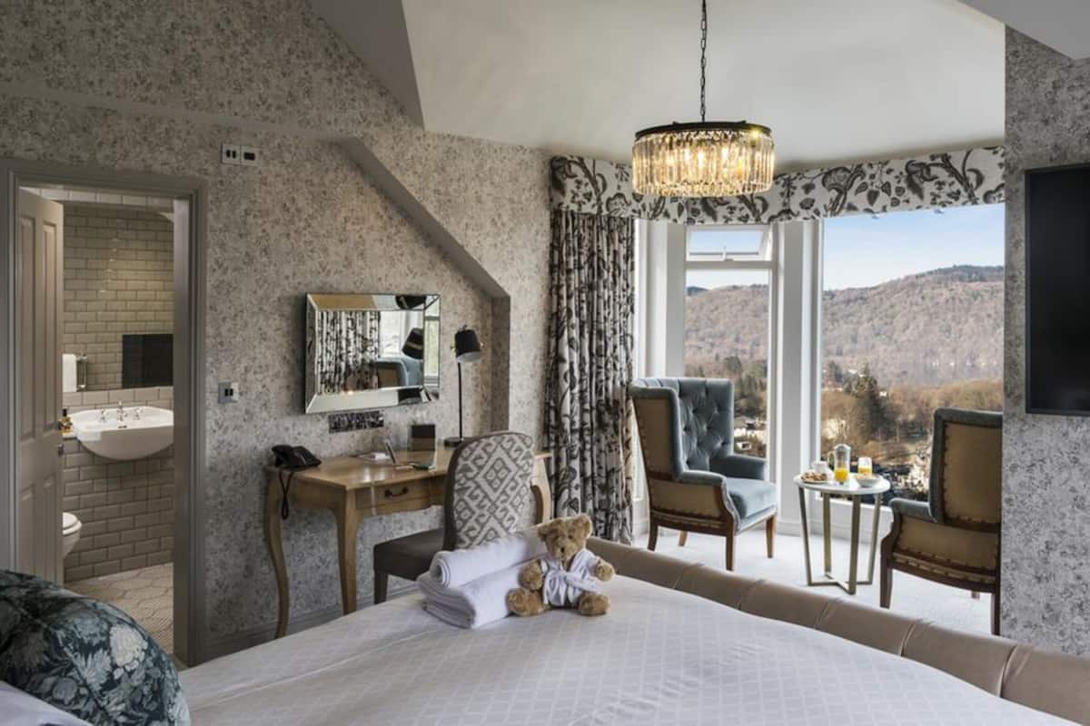 A bedroom with a large bed, a desk, a chair, a small table with drinks, a teddy bear, a mirror, and a view of the mountains.