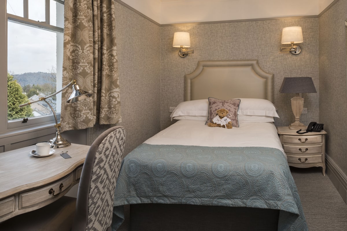 A neatly arranged hotel room with a bed, bedside lamps, a desk with a lamp, a chair, and a view of the outdoors.