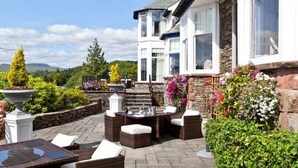 An outdoor patio area with wicker furniture, a stone wall, and a view of a two-story building with large windows and a stone facade. - Windermere Hillthwaite Hotel (Windermere)