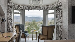 A hotel room with a large window, a desk, two chairs, a round table with breakfast, and a bed. - Windermere Hillthwaite Hotel (Windermere)