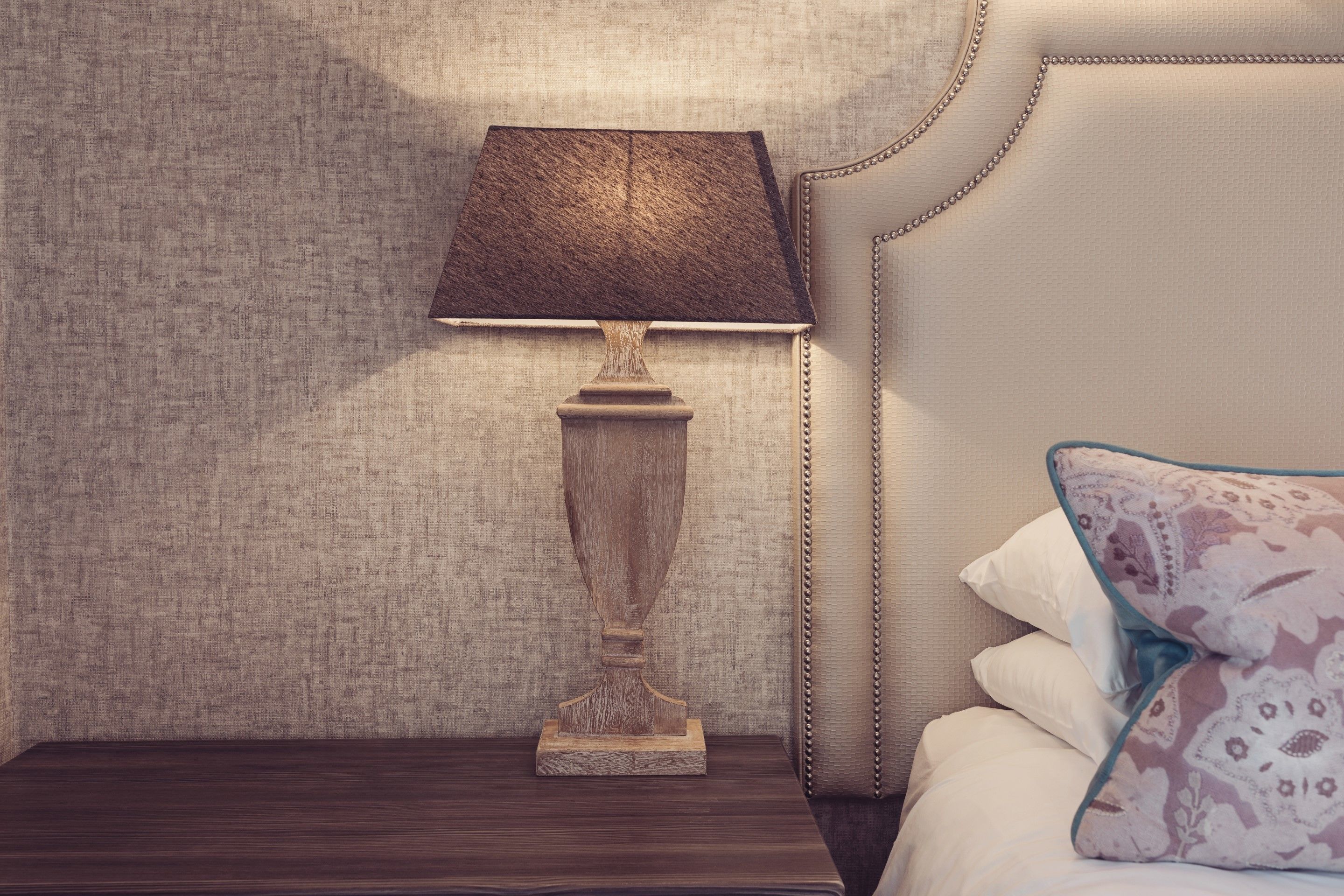 Una mesita de noche de madera con una lámpara, una cama con cabecero y un cojín estampado.