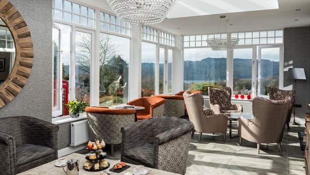 A sunlit room with large windows, patterned furniture, and a chandelier. - Windermere Hillthwaite Hotel (Windermere)