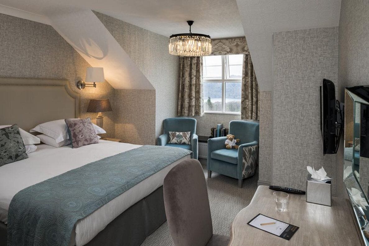 Ein Hotelzimmer mit einem Bett, zwei Sesseln, einem Schreibtisch mit Lampe, einem Fernseher und einem Fenster mit Vorhängen.