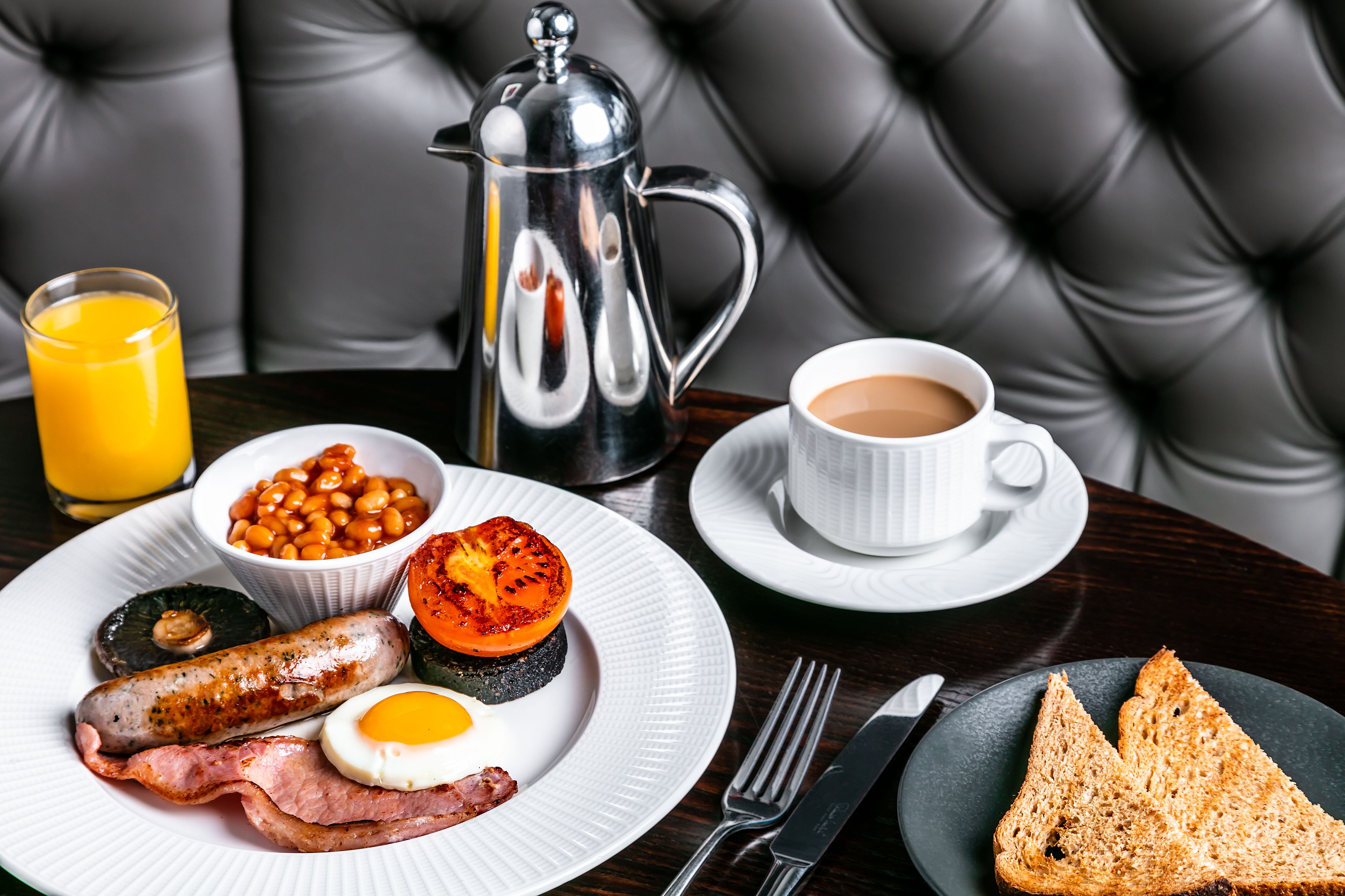A breakfast setting with a plate of eggs, bacon, sausages, beans, and toast, a cup of coffee, and a glass of orange juice on a table with a black leather couch in the background.