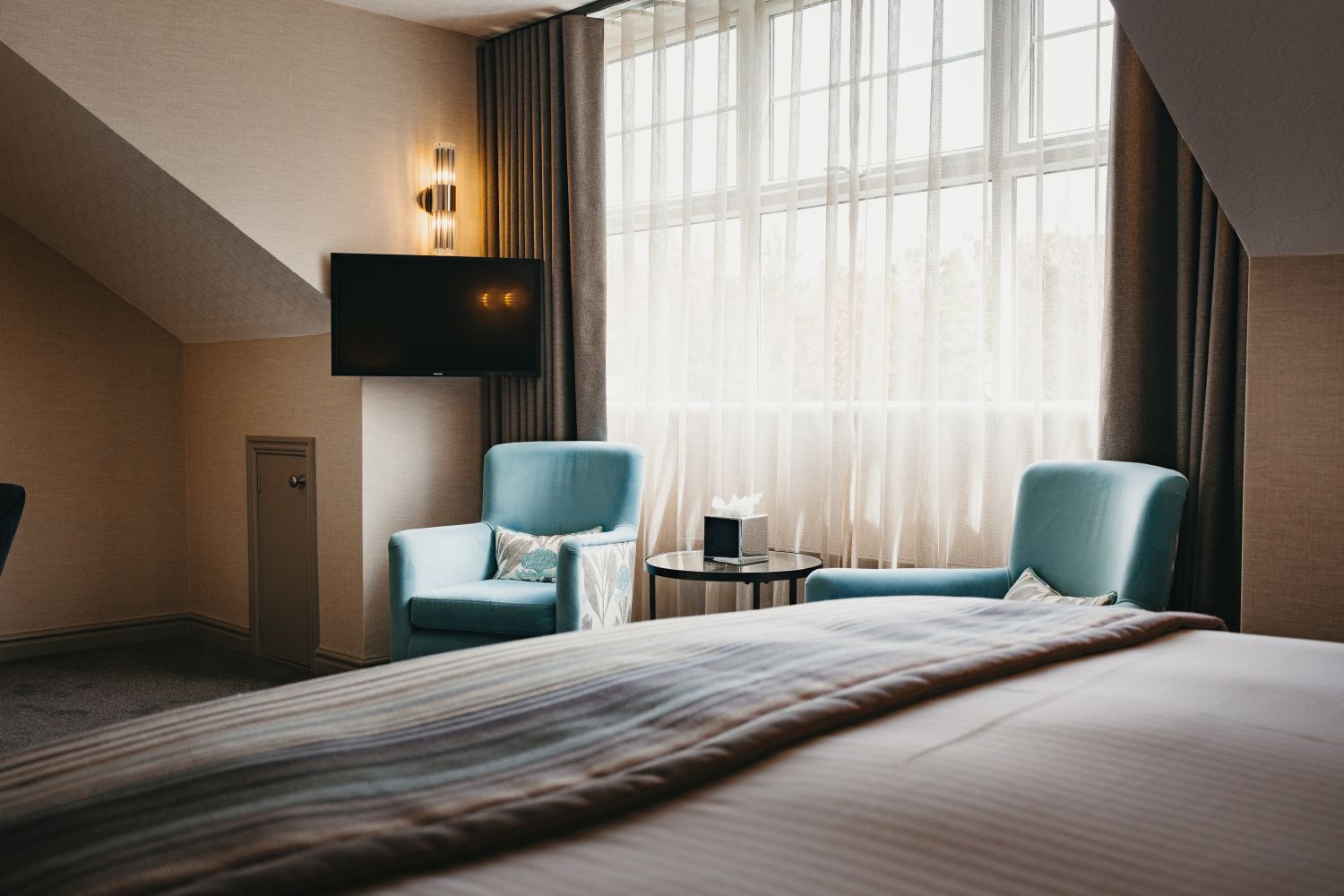 A hotel room with a large bed, two armchairs, a small round table with a lamp, a TV mounted on the wall, and a window with sheer curtains.