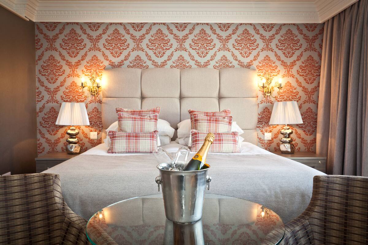 A hotel room with a bed, two nightstands with lamps, a glass table with a bottle of champagne, and two upholstered chairs.