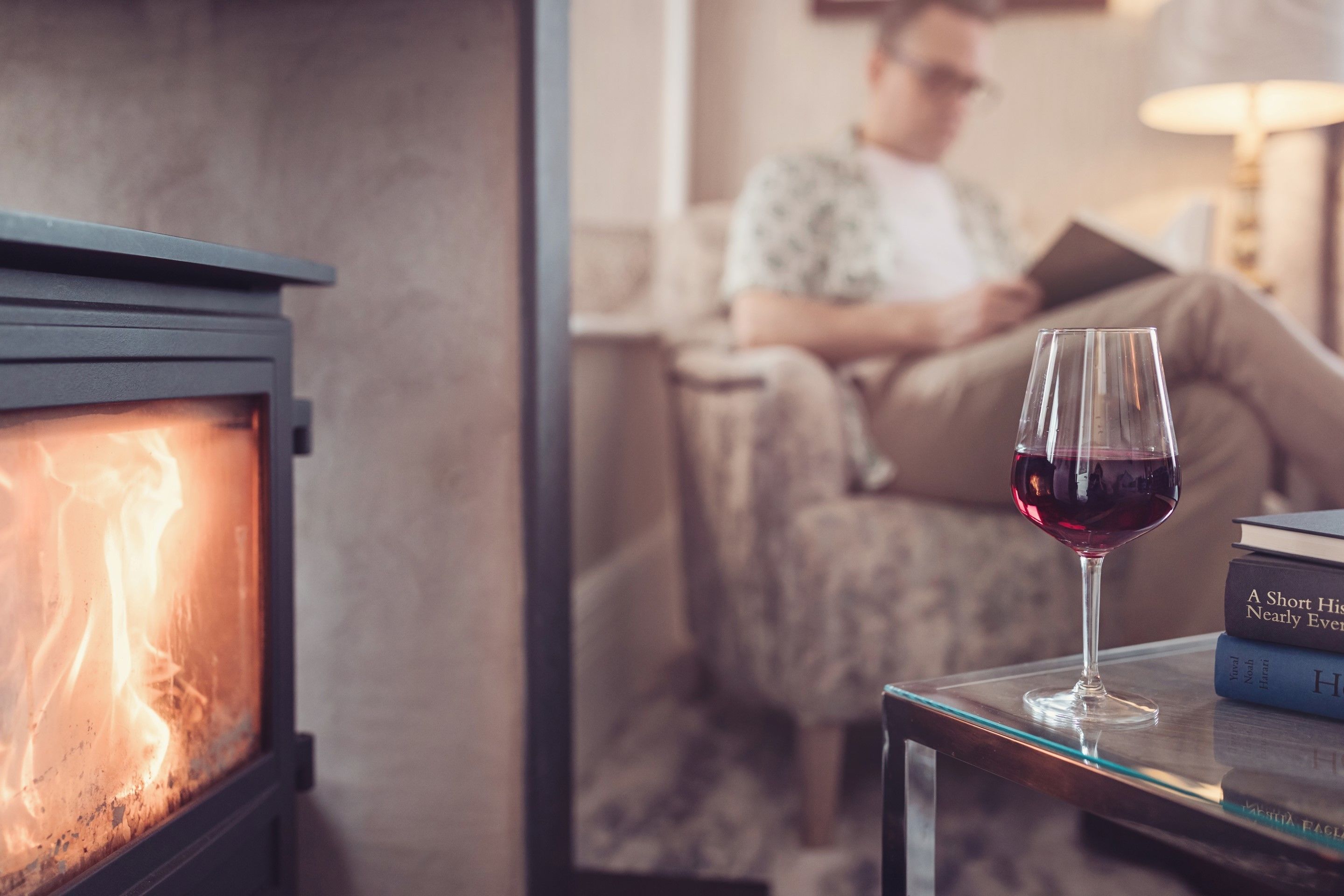 A cozy living room with a fireplace, a glass table with a wine glass, and a person reading a book.