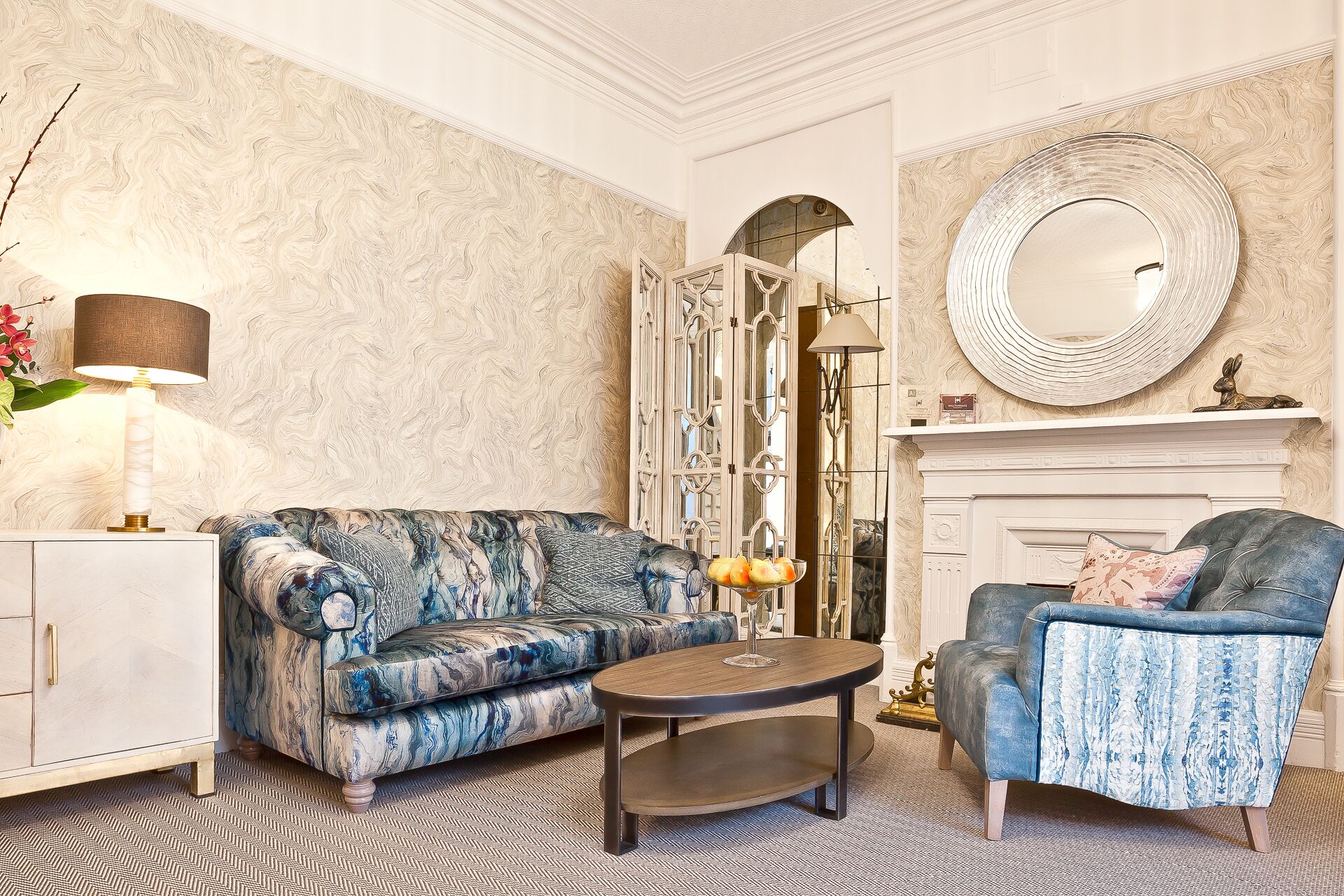 Un salon avec un canapé à motifs, une table basse ronde, un fauteuil bleu, une cheminée et un miroir décoratif.