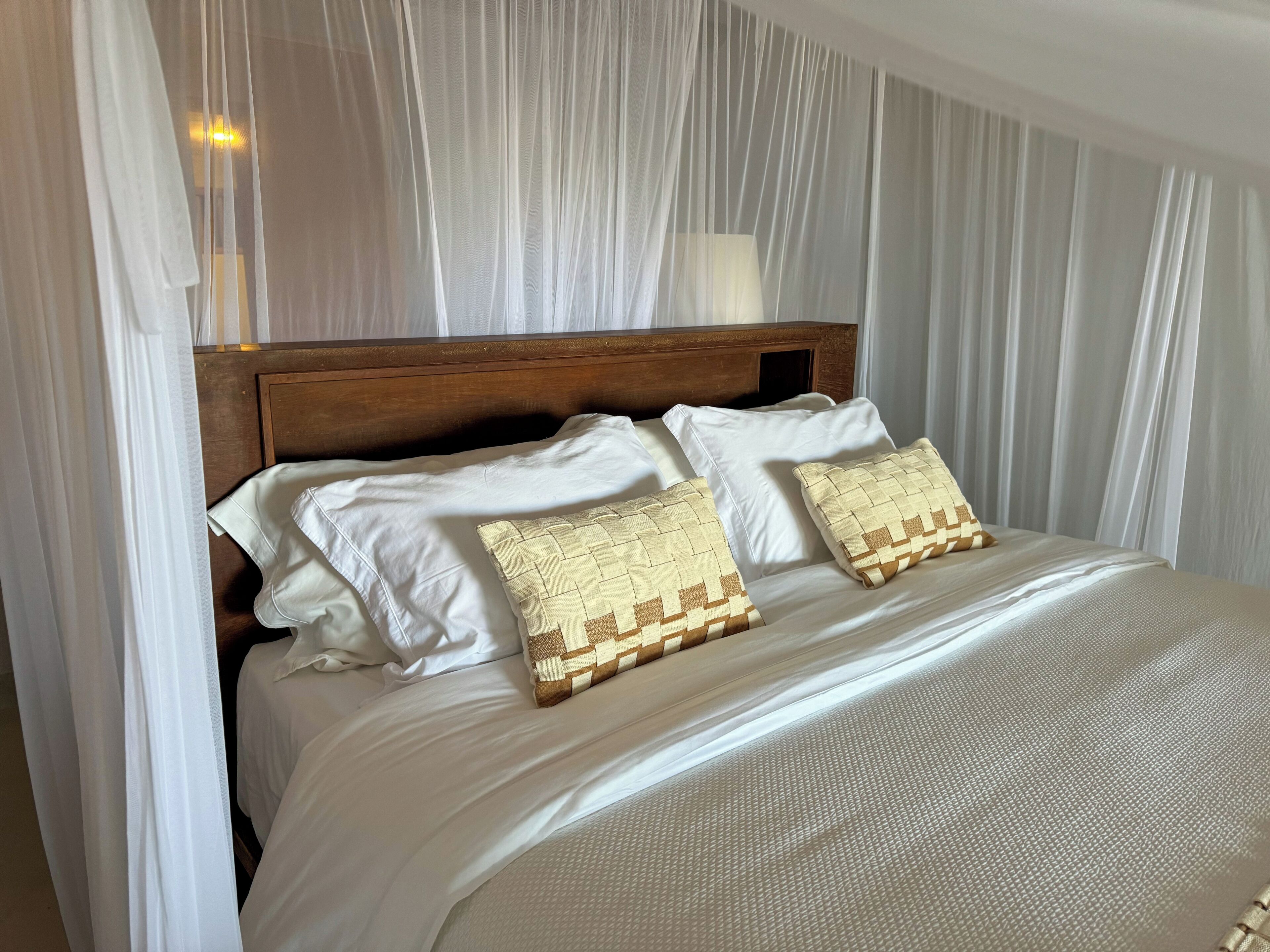 A neatly made bed with white linens and two patterned pillows, a wooden headboard, and sheer white curtains.