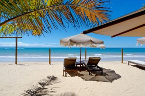 Une plage avec deux chaises longues sous un parasol à rayures, un palmier et l’océan en arrière-plan.