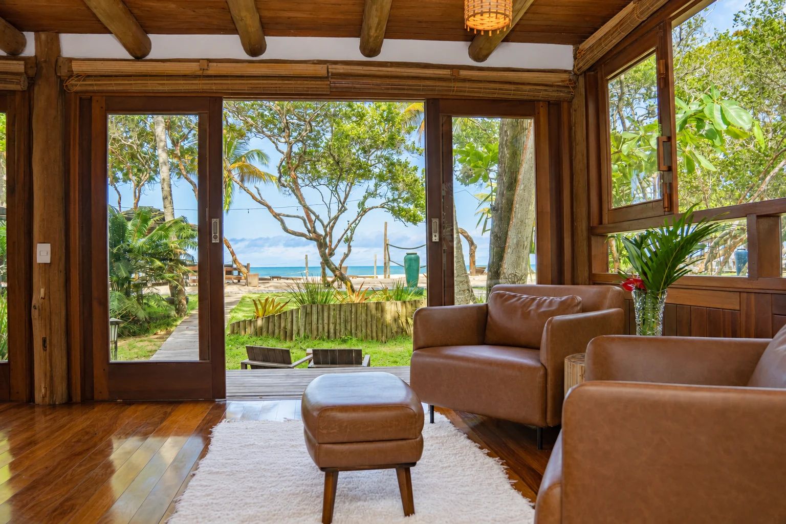 Quarto com piso de madeira, sofá de couro marrom, tapete branco e vista para uma praia através de grandes portas de vidro.