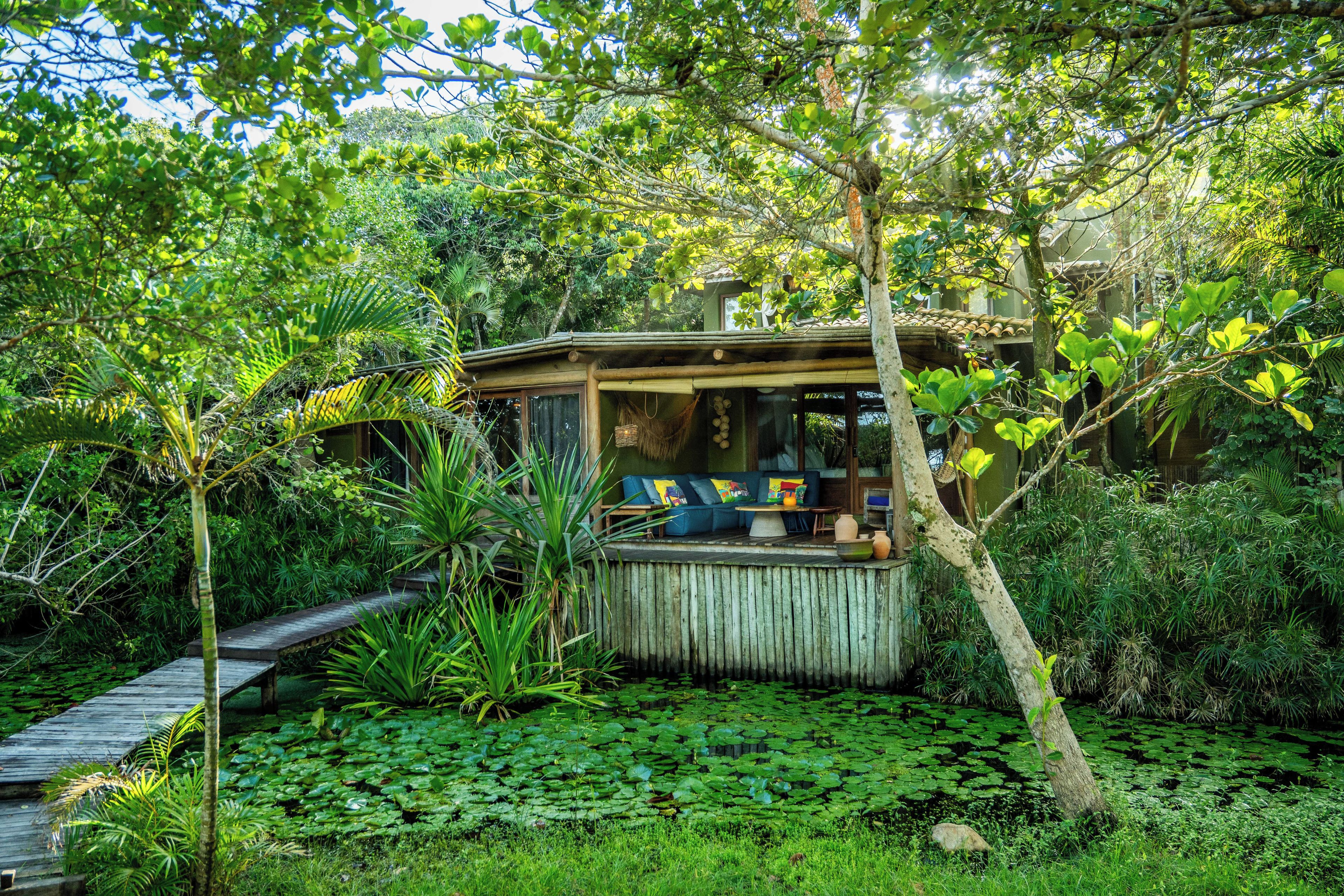 Uma cabana de madeira com varanda, cercada por vegetação exuberante e um lago com nenúfares.