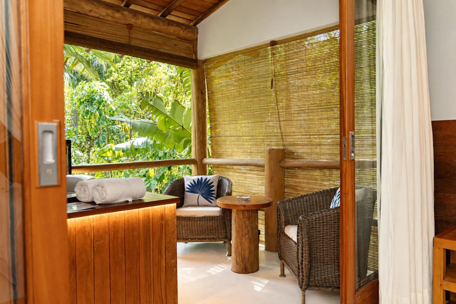 Quarto com teto de madeira, porta de vidro para uma varanda, móveis de vime e vista para vegetação exuberante.
