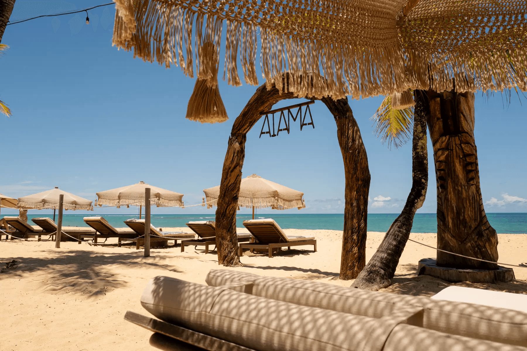 Uma praia com cadeiras de praia e guarda-sóis, um letreiro com a inscrição 'AIAMA', e um céu azul e límpido.