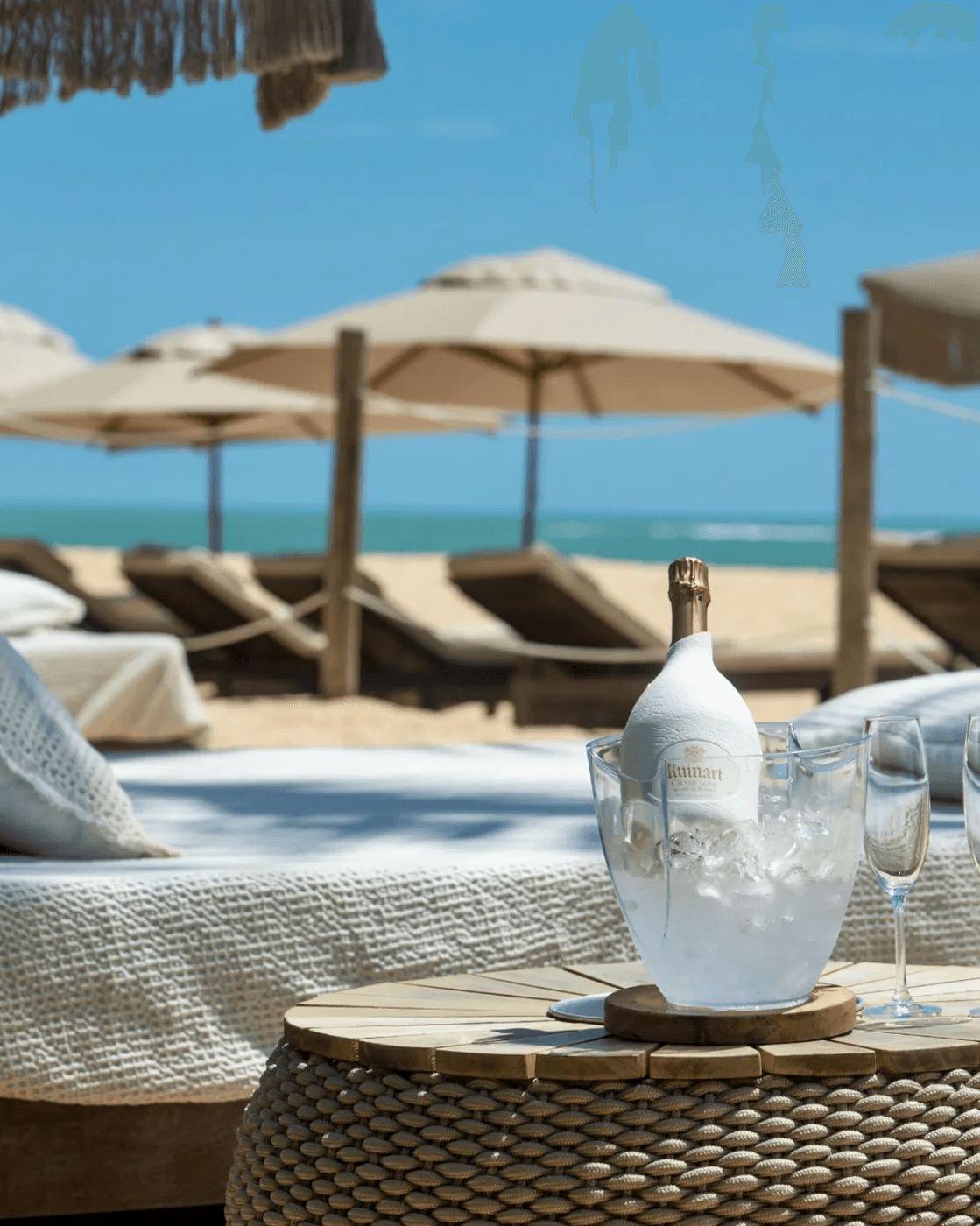Uma área de estar à beira-mar com uma garrafa de champanhe Ruinart numa taça, duas taças de champanhe e cadeiras de lounge sob guarda-sóis.