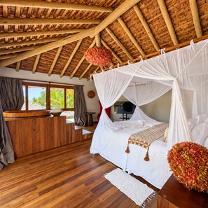 A room with a wooden ceiling, a bed with white bedding, a wooden desk, and a large window.