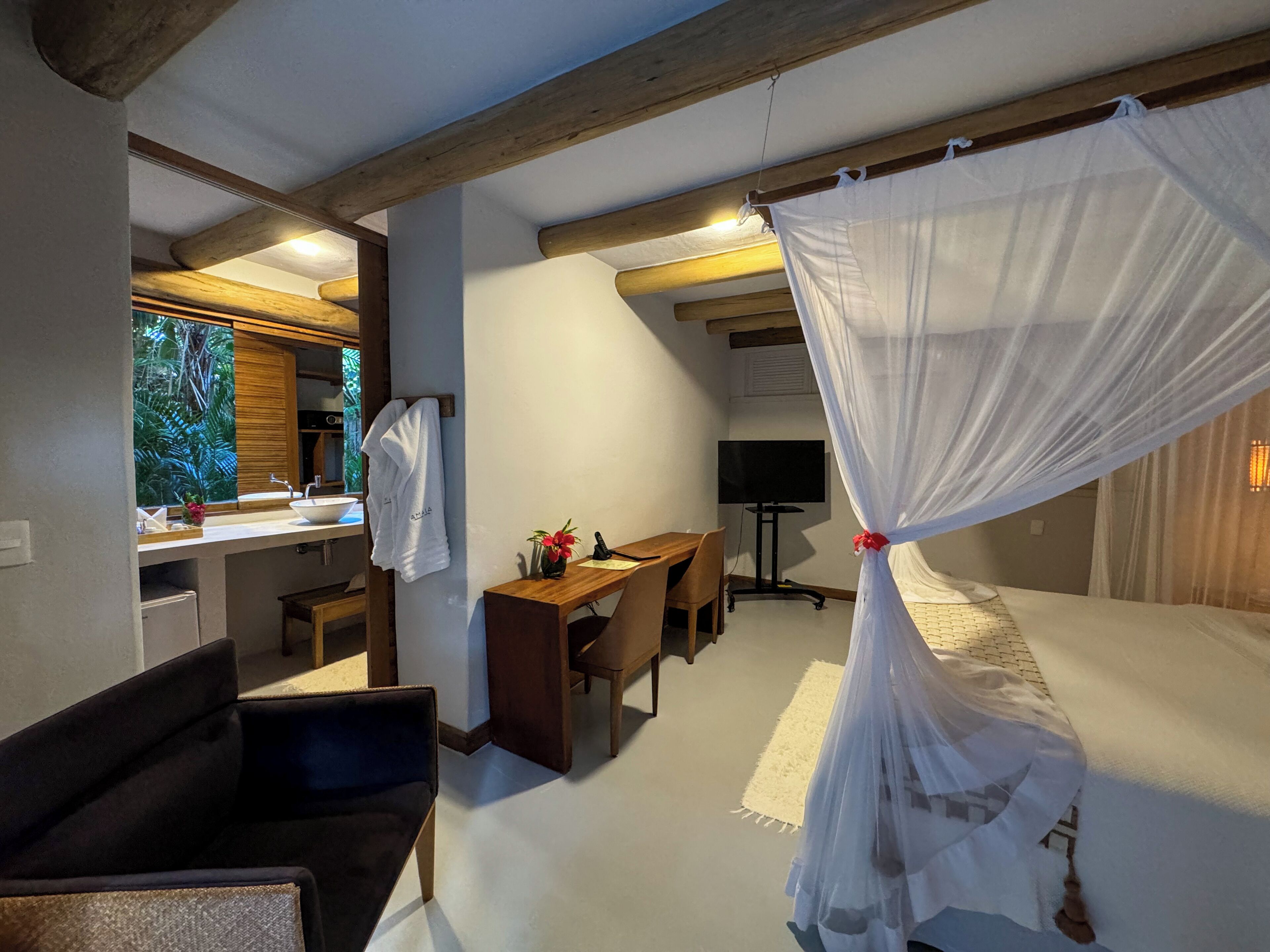A bedroom with a canopy bed, a desk with a chair, a TV, and a bathroom with a sink and mirror.