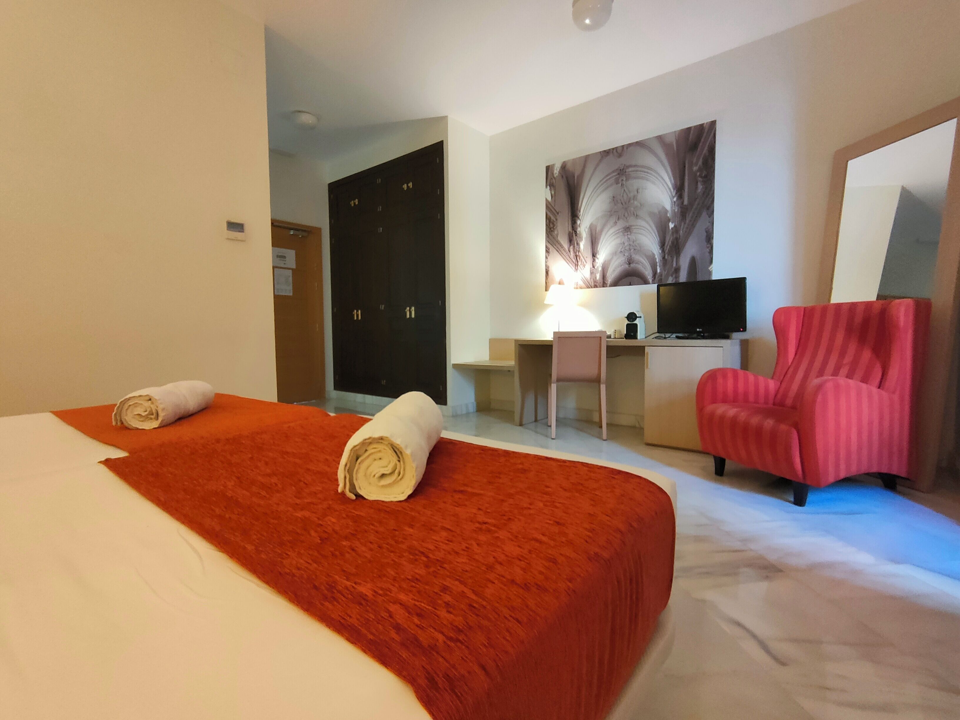 A hotel room with a large bed, a desk with a computer, a TV, and a red chair.