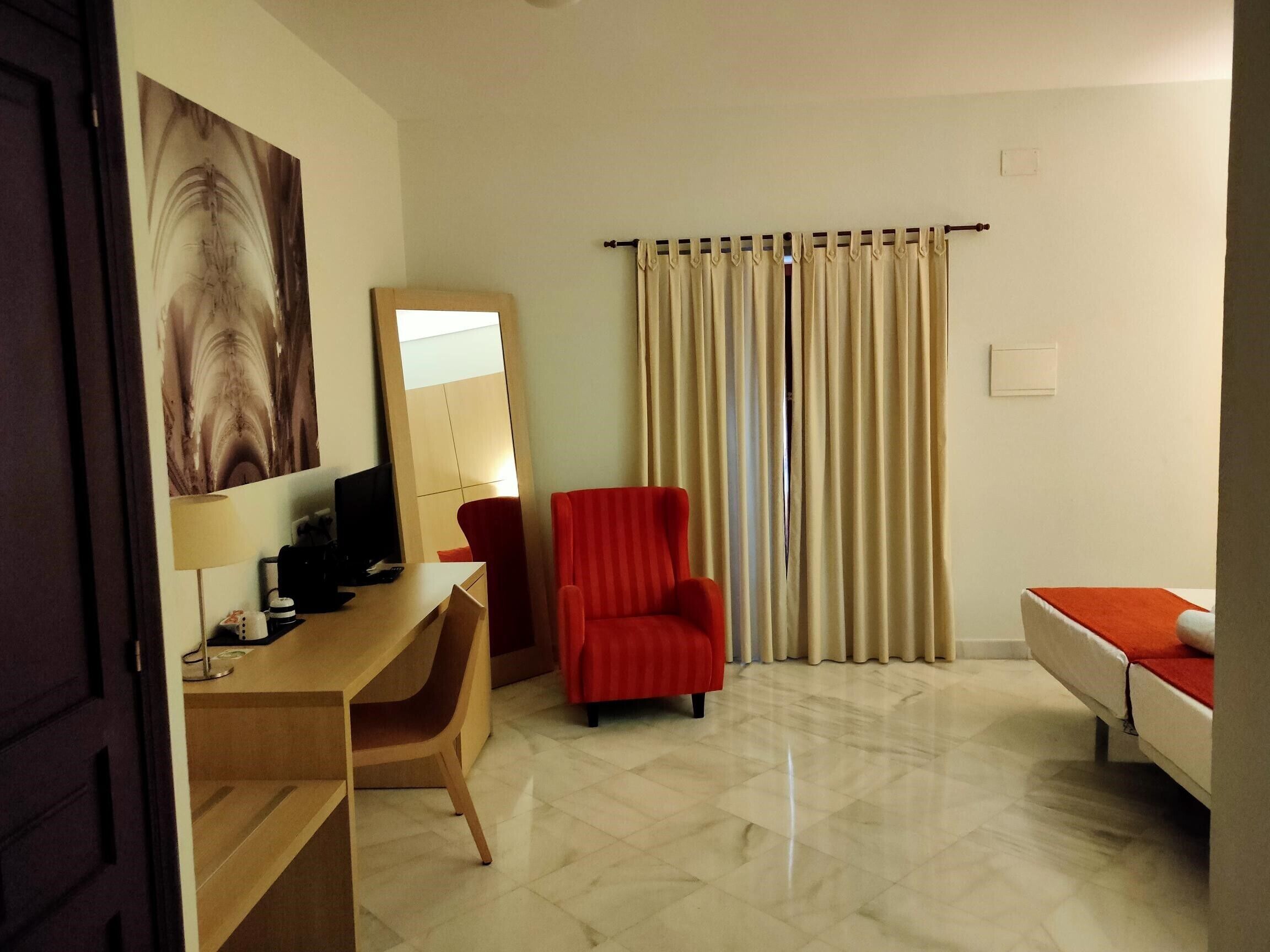 A hotel room with a desk, a red armchair, a bed with an orange bedspread, and a large window with beige curtains.