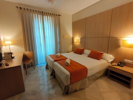 Habitación de hotel con cama doble, dos mesitas de noche, un escritorio con silla, una lámpara y una ventana con cortinas.