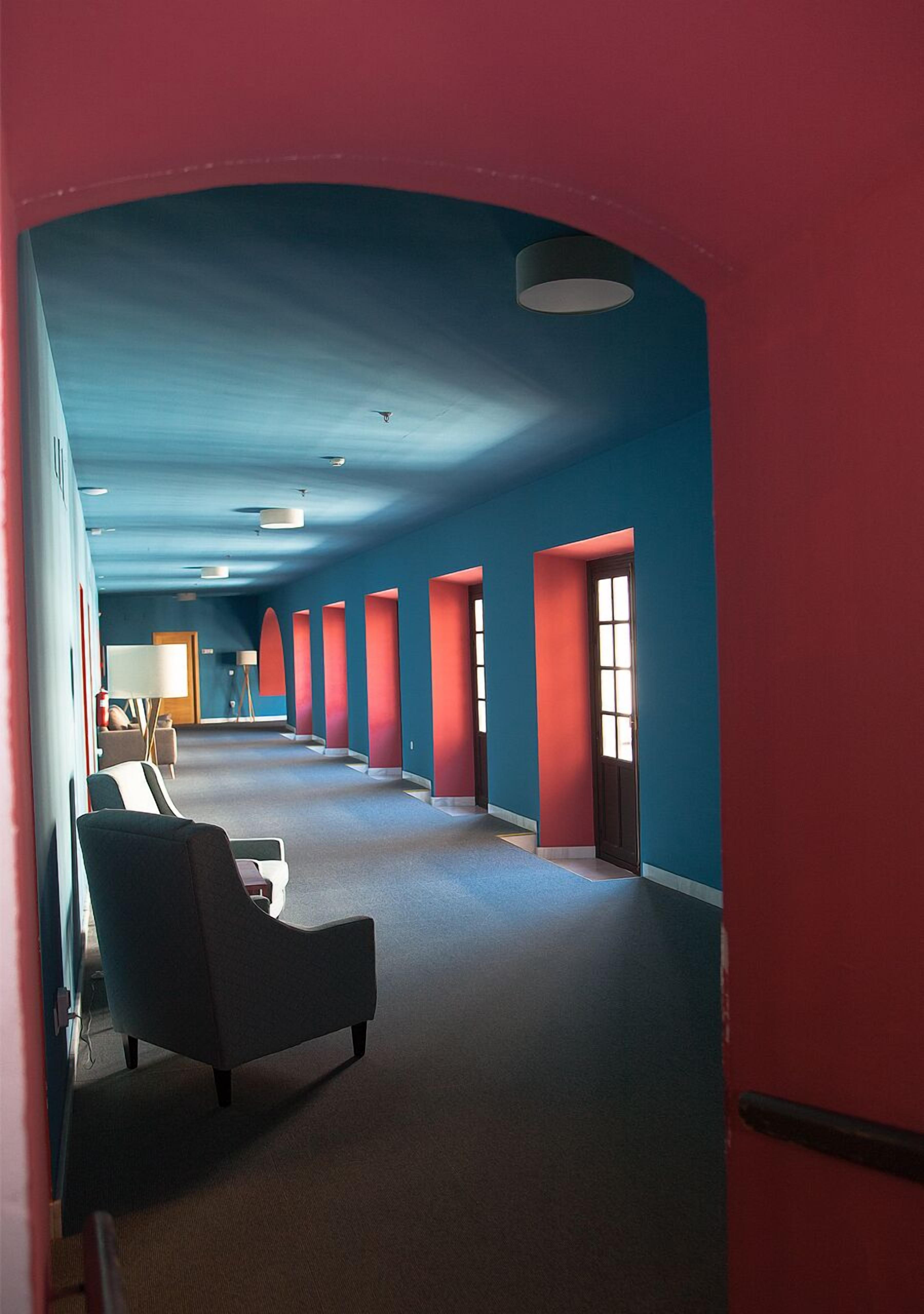 A hallway with red and blue walls, a row of doors, and a seating area with chairs.