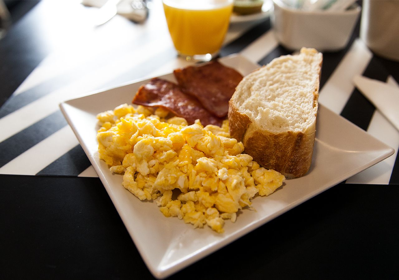 A breakfast plate with scrambled eggs, bacon, toast, and a glass of orange juice on a striped tablecloth.