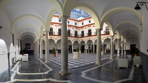 A spacious courtyard with a checkered floor, white columns, and a building with red-tiled roofs in the background. - Hotel Boutique Convento Cádiz (Cádiz)