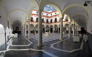 Un patio espacioso con piso a cuadros, columnas blancas y un edificio con tejados rojos al fondo.