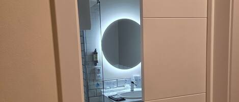 Un baño moderno con cabina de ducha de vidrio, un espejo redondo y un lavamanos blanco.