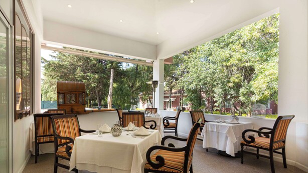 A restaurant with tables set for dining, surrounded by chairs, and large windows offering a view of trees. - MM Grand Hotel Puebla, Tapestry Collection by Hilton (Puebla)