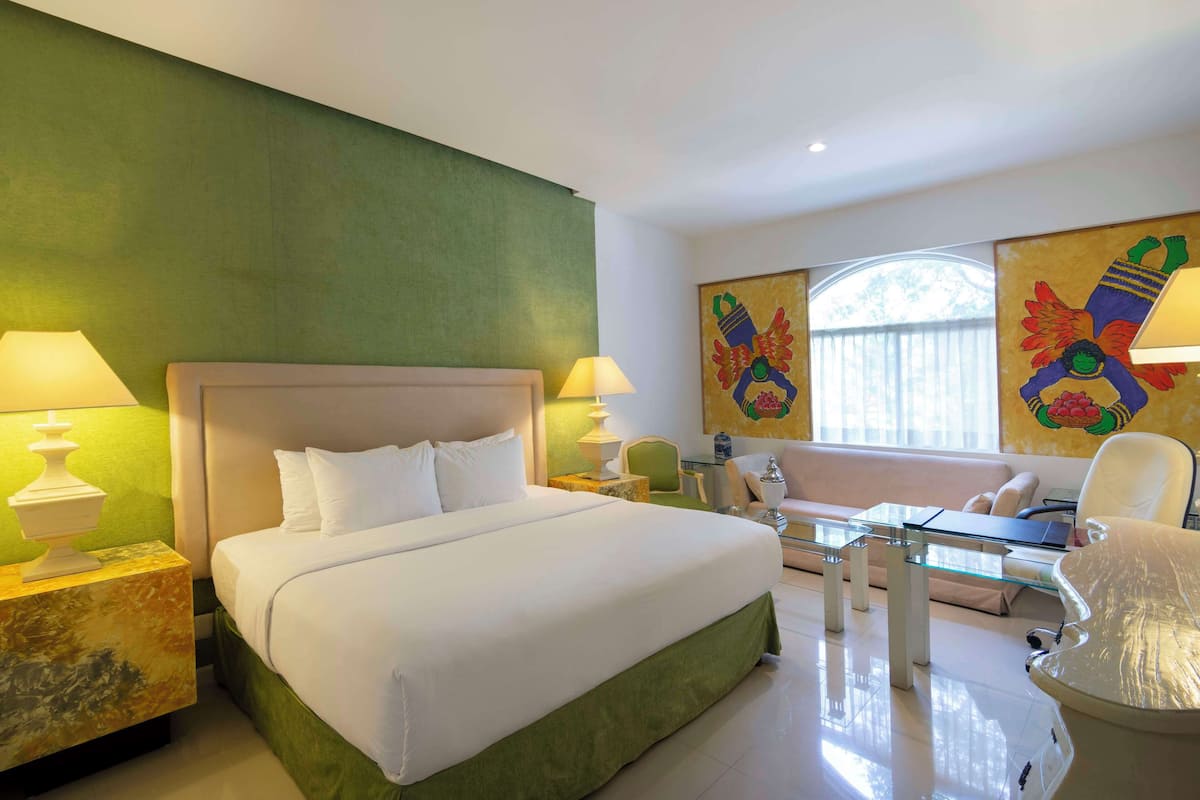 A hotel room with a large bed, a glass coffee table, a sofa, and two chairs. There are two lamps on the nightstands.