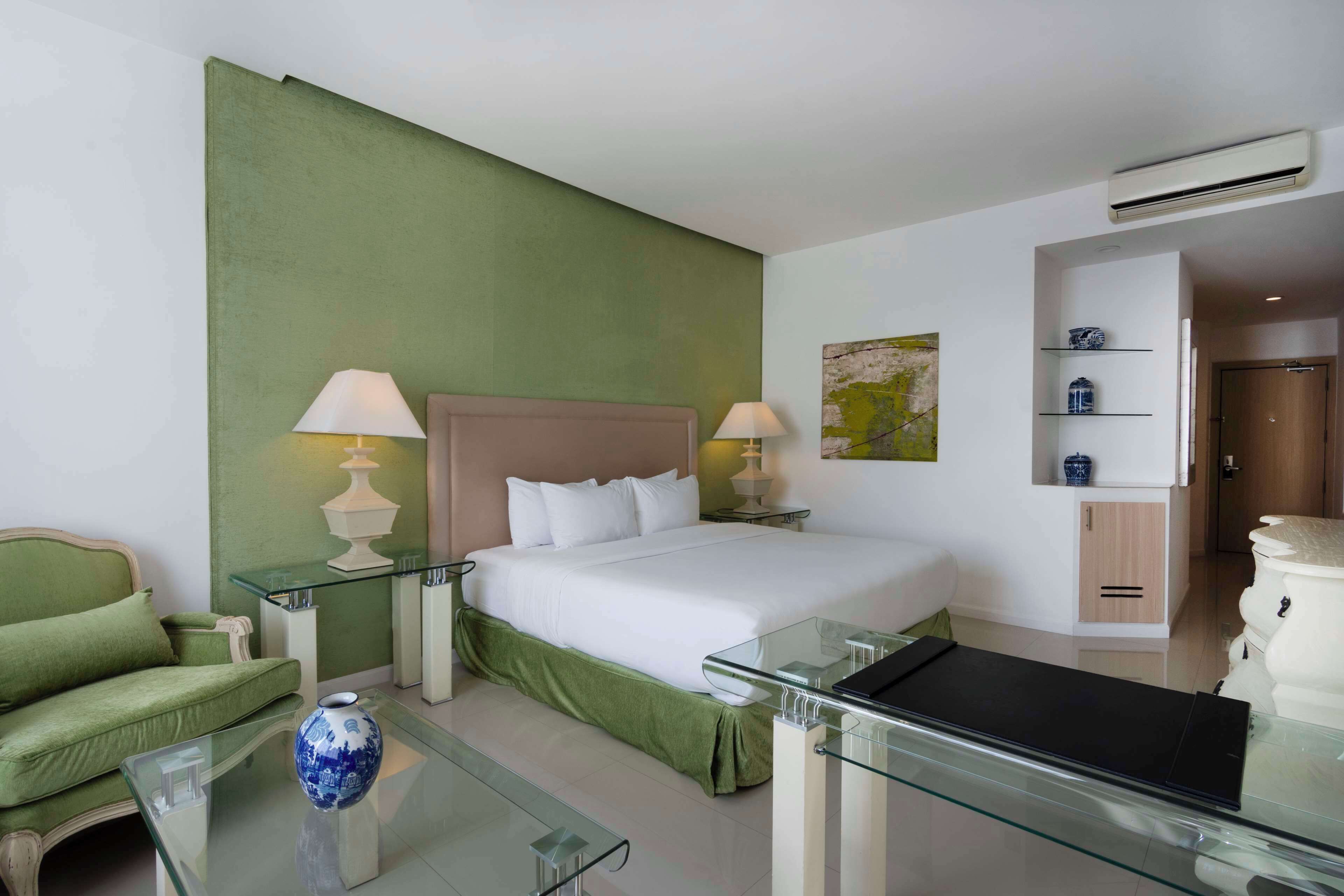 A hotel room with a bed, green walls, a glass coffee table, a green sofa, and a vase.