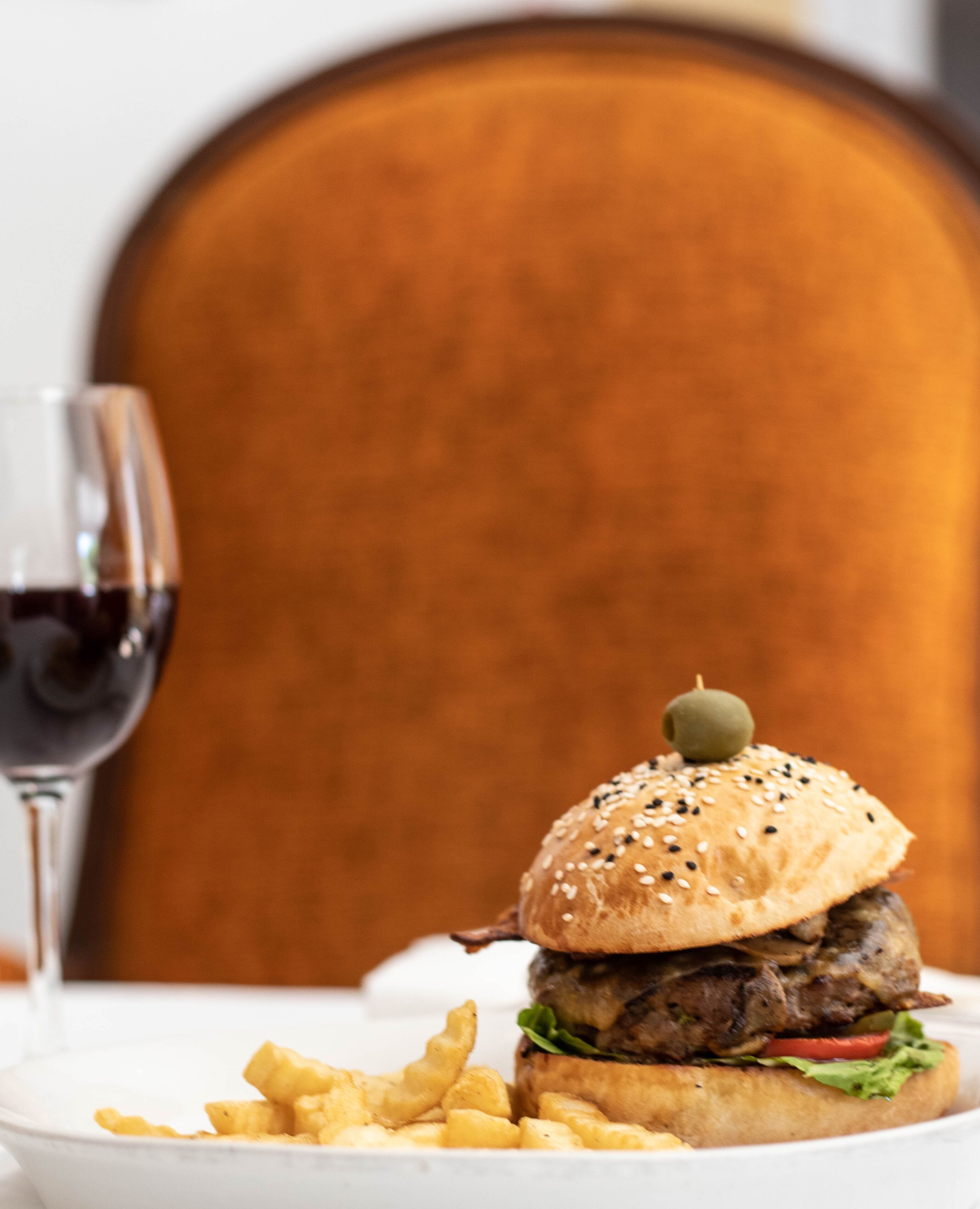 A burger with a sesame seed bun and an olive on top, served with fries on a white plate.