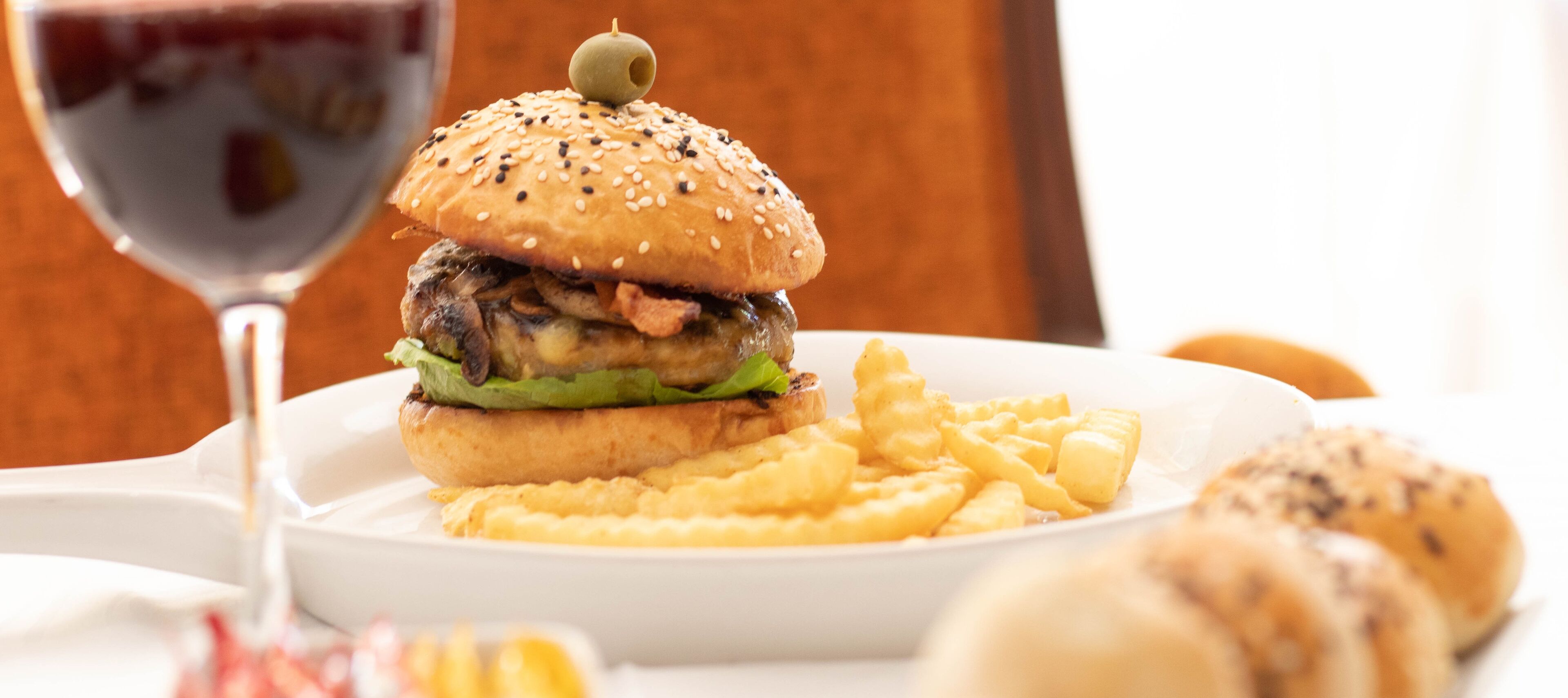 Ein Burger mit Sesambrötchen und grüner Garnitur, serviert mit Pommes und einem Glas Rotwein.