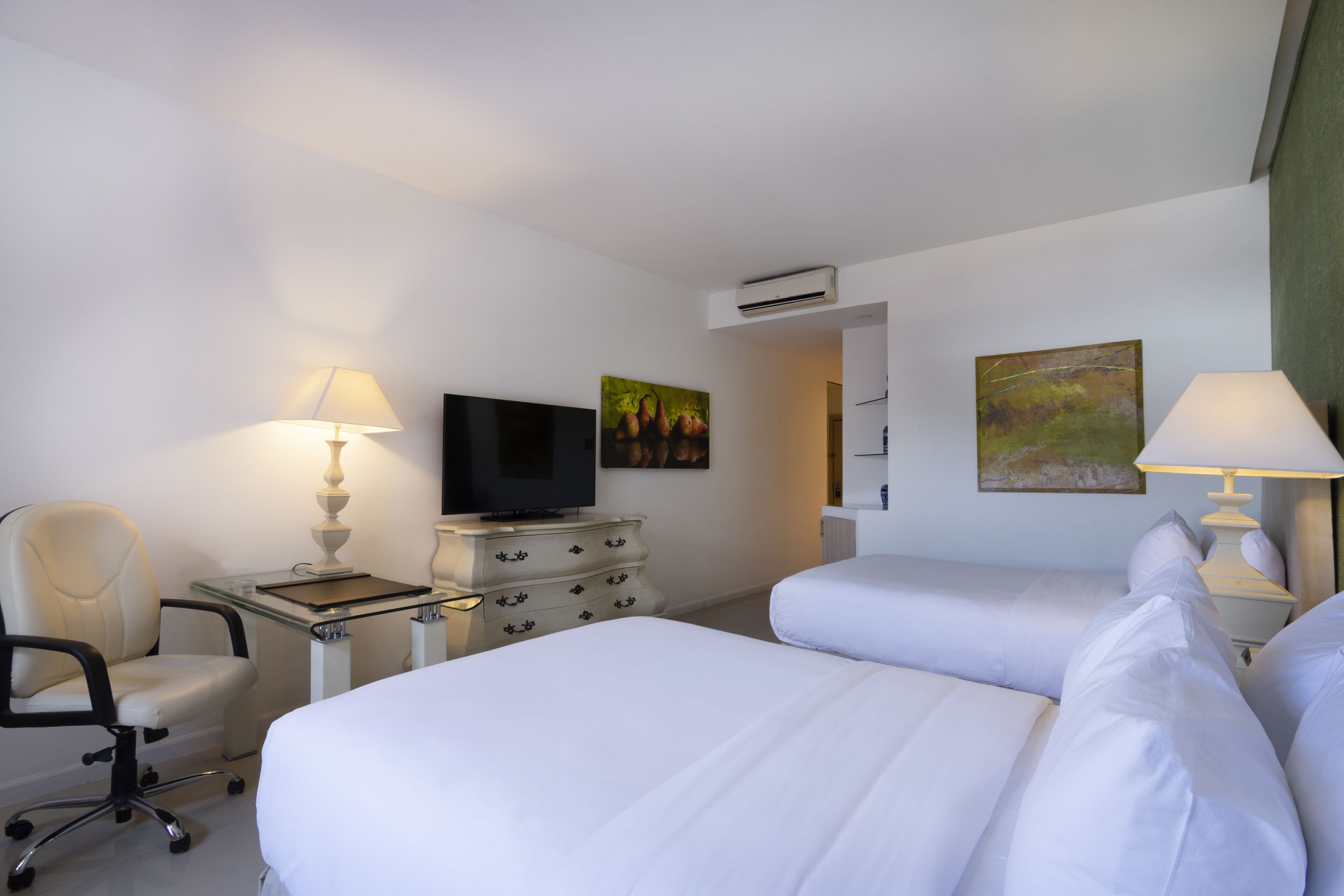 A hotel room with two beds, a desk with a lamp, a chair, a television, and artwork on the walls.
