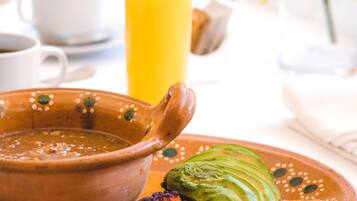 A breakfast setting with a clay bowl, sliced avocado, grilled meat, and a glass of orange juice.