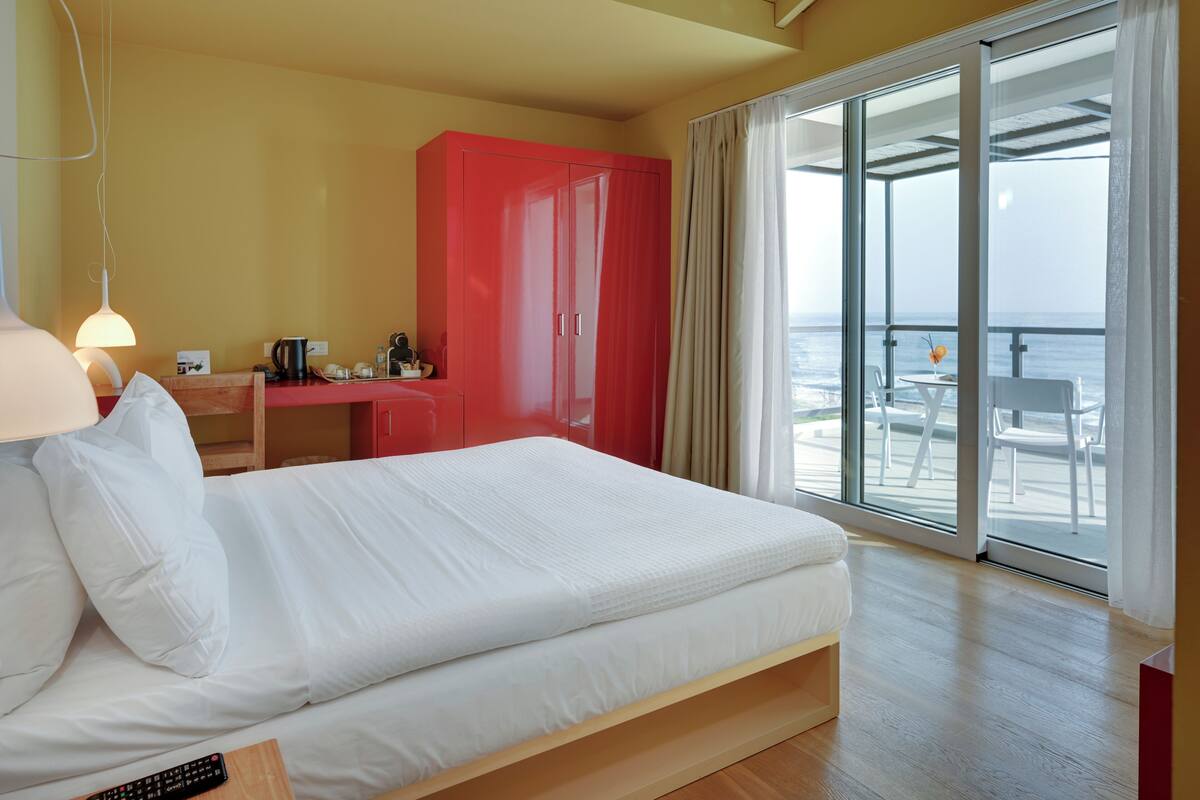 A hotel room with a bed, a desk, a red wardrobe, and a balcony with a view of the sea.