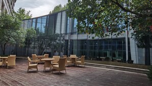 An outdoor seating area with wicker furniture and a wooden deck, surrounded by trees and a modern building with large glass windows. - Sheraton Qinhuangdao Beidaihe Hotel (Qinhuangdao)