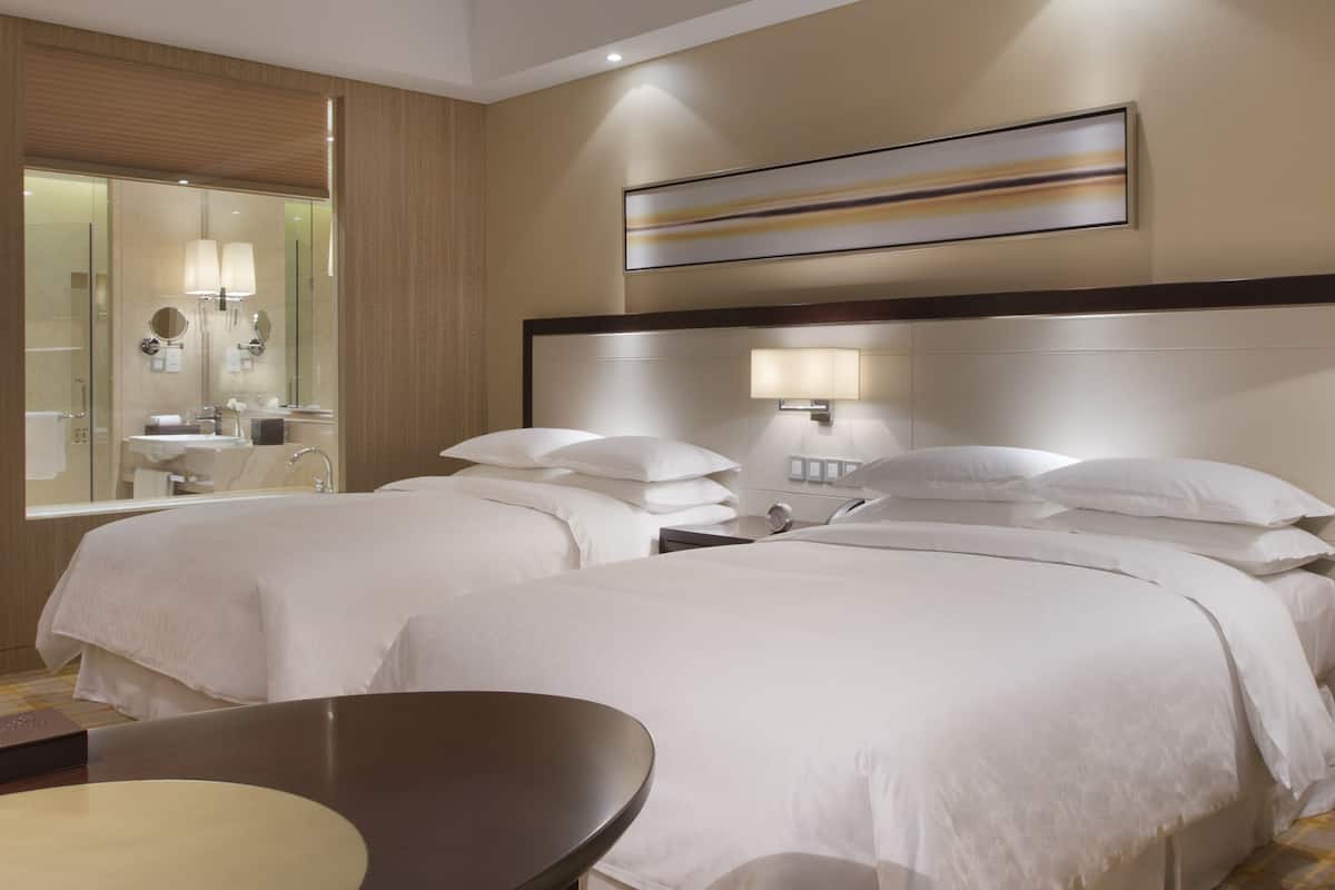 Une chambre d’hôtel avec deux lits, une table ronde, un grand miroir et une œuvre d’art moderne accrochée au mur.