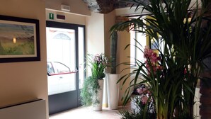 Un hall d’hôtel avec un sol carrelé, un tableau représentant un paysage encadré, des plantes en pot et une porte donnant sur l’extérieur.