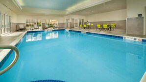 Une piscine intérieure à l’eau claire, avec des bords carrelés et un escalier pour y accéder. Des chaises longues et des tables sont disposées autour de la piscine.