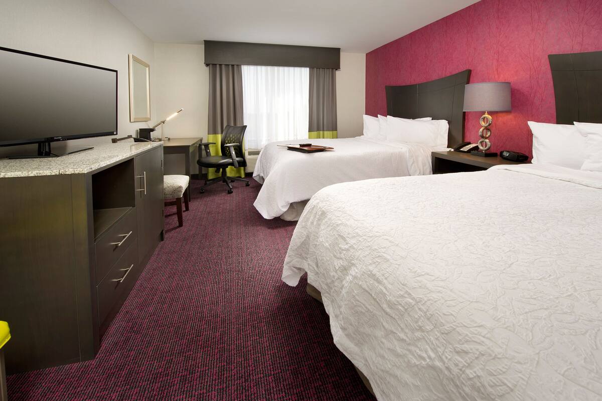 Ett hotellrum med två sängar, ett skrivbord med en platt-TV, en stol, en lampa och ett nattduksbord med en telefon.