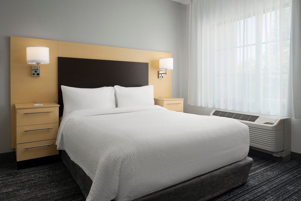 Een hotelkamer met een groot bed, twee nachtkastjes, een raam met dunne gordijnen en een wandairconditioning.