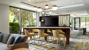 Un hall d’hôtel moderne doté d’un grand espace salon, d’un bar avec des tabourets, d’un téléviseur à écran plat et de grandes fenêtres offrant une vue sur l’extérieur.