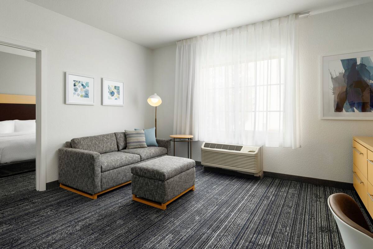 Een hotelkamer met een grijze bank, een poef, een klein rond tafeltje, een staande lamp, een bed, een witte airconditioning en een houten dressoir.