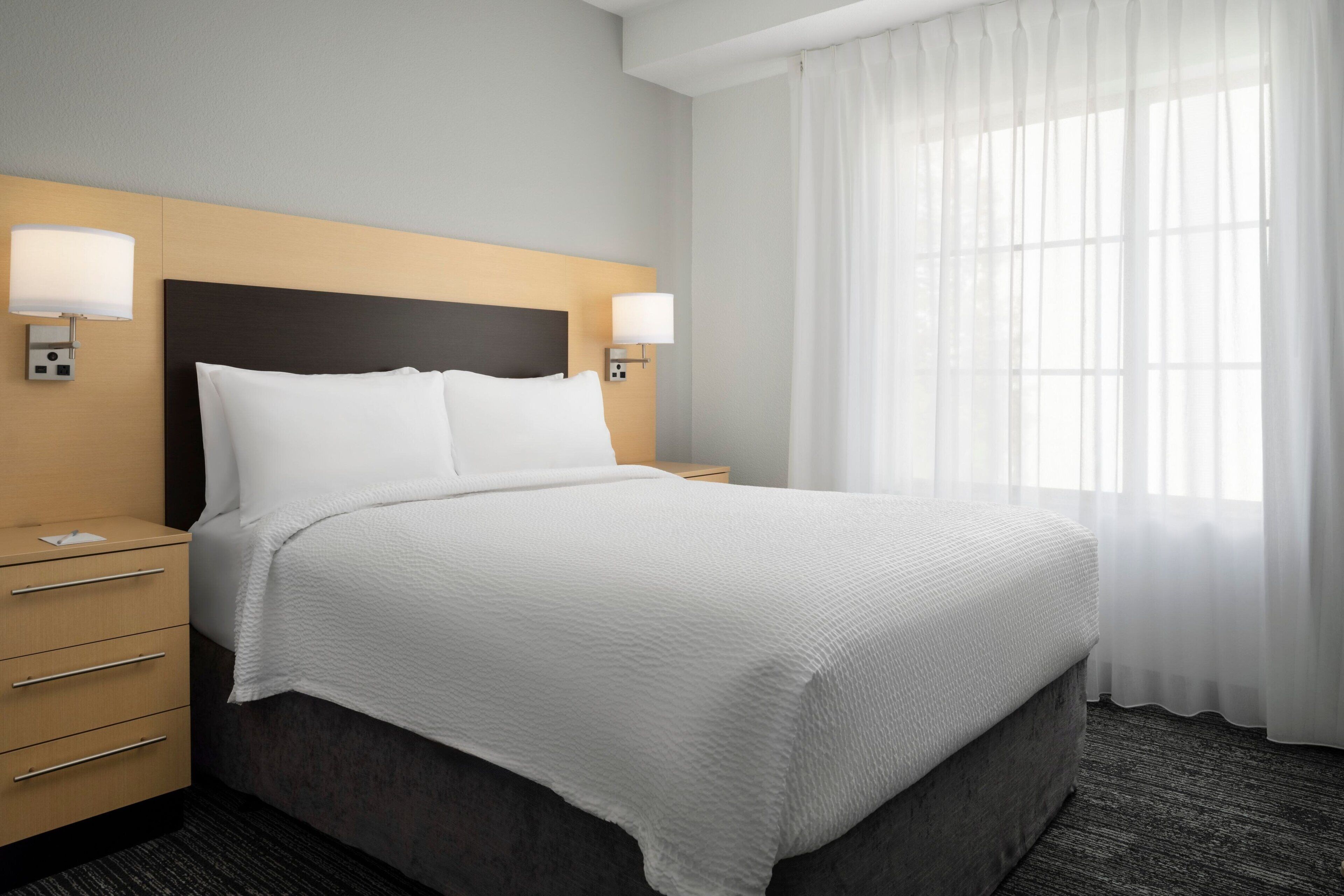 Habitación de hotel con una cama grande, dos mesitas de noche, un cabecero de madera y una ventana con cortinas translúcidas.